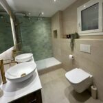 Presentamos una transformación radical ejecutada por Reformas Guimerà. Descubre cómo renovamos un baño anticuado sustituyendo el mármol y la bañera por una ducha a ras de suelo, apostando por el blanco puro y la calidez de la madera cerámica para ganar amplitud y confort.