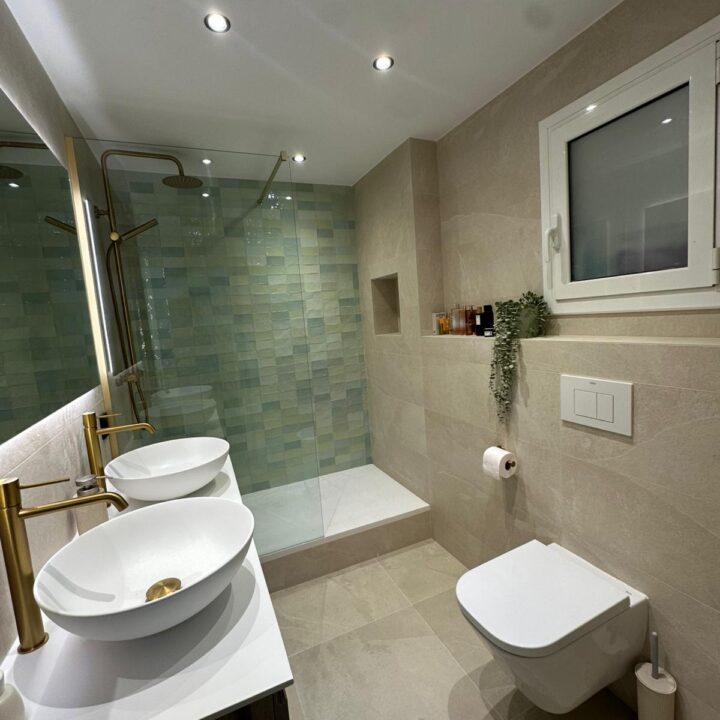 Presentamos una transformación radical ejecutada por Reformas Guimerà. Descubre cómo renovamos un baño anticuado sustituyendo el mármol y la bañera por una ducha a ras de suelo, apostando por el blanco puro y la calidez de la madera cerámica para ganar amplitud y confort.