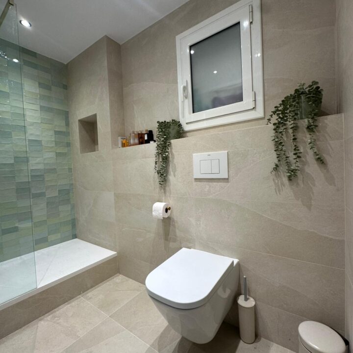 Presentamos una transformación radical ejecutada por Reformas Guimerà. Descubre cómo renovamos un baño anticuado sustituyendo el mármol y la bañera por una ducha a ras de suelo, apostando por el blanco puro y la calidez de la madera cerámica para ganar amplitud y confort.